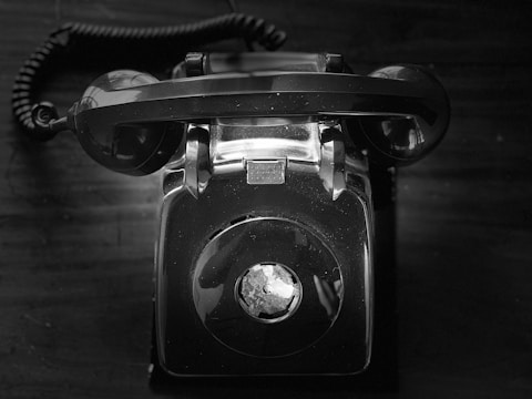 A vintage rotary telephone is centered in the image, sitting on a dark surface. The phone is black and white, with a coiled cord visible in the top left corner. The rotary dial and the handset cradle are prominently displayed, indicating a classic design.