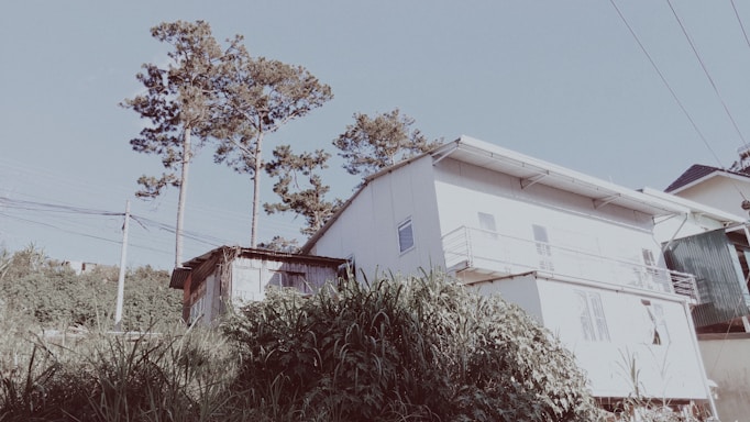 A modest residential building with a white facade and a small balcony, surrounded by tall grass and trees. The sky is clear with visible power lines in the foreground.