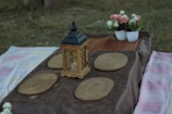 A picnic setup on a grassy area features a wooden table with a dark tablecloth, adorned with a decorative lantern at the center and surrounded by several woven placemats. In the background, there are two white pots filled with pink and white flowers.