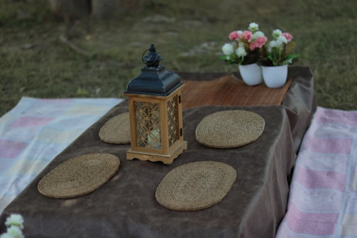 A picnic setup on a grassy area features a wooden table with a dark tablecloth, adorned with a decorative lantern at the center and surrounded by several woven placemats. In the background, there are two white pots filled with pink and white flowers.