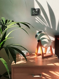 A wooden table features a stylish lamp with a wooden stand casting soft shadows. Lush green leaves of a plant are visible, creating intricate patterns on the wall. The scene has a serene atmosphere with sunlight streaming in, highlighting the natural elements.