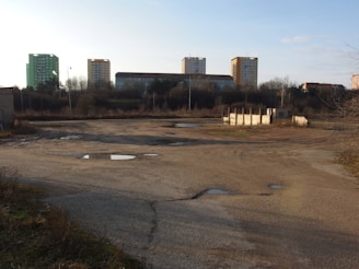 An open and barren landscape with patches of water on a rough surface, surrounded by overgrown vegetation. In the background are several tall residential buildings with a mix of green and beige colors. The sky is clear.