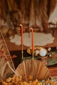 Two tall, red candles are lit and placed in ornate holders on a dark wooden surface, accompanied by a white round candle. The setting includes dried palm leaves, yellow flowers, and a blurred background with earthy tones and a rustic style.