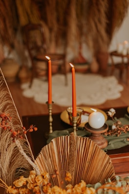 Two tall, red candles are lit and placed in ornate holders on a dark wooden surface, accompanied by a white round candle. The setting includes dried palm leaves, yellow flowers, and a blurred background with earthy tones and a rustic style.