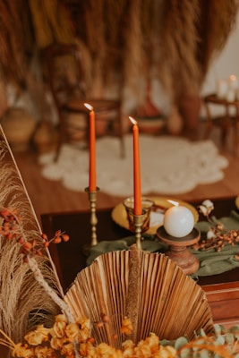 Two tall, red candles are lit and placed in ornate holders on a dark wooden surface, accompanied by a white round candle. The setting includes dried palm leaves, yellow flowers, and a blurred background with earthy tones and a rustic style.
