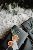 Two neatly wrapped gifts adorned with dried orange slices and evergreen branches, set against a textured white surface and partially under a fir tree.