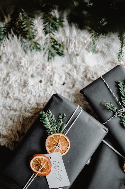 Two neatly wrapped gifts adorned with dried orange slices and evergreen branches, set against a textured white surface and partially under a fir tree.