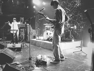 A black and white scene of a live music performance on a stage with musicians playing instruments. The stage is equipped with various musical and audio equipment, including microphones, speakers, and amplifiers. One person is playing an electric guitar, while another stands near a keyboard. The background features a set of drums and stage lighting, creating an energetic and vibrant atmosphere.