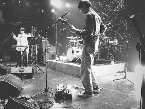 A black and white scene of a live music performance on a stage with musicians playing instruments. The stage is equipped with various musical and audio equipment, including microphones, speakers, and amplifiers. One person is playing an electric guitar, while another stands near a keyboard. The background features a set of drums and stage lighting, creating an energetic and vibrant atmosphere.