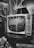A vintage television set with dials sits on a table surrounded by patterned boxes. The reflection of nearby objects can be seen on the TV screen, and a jacket is visible next to it. The setting has a nostalgic, retro feel with a mix of geometric patterns.