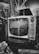 A vintage television set with dials sits on a table surrounded by patterned boxes. The reflection of nearby objects can be seen on the TV screen, and a jacket is visible next to it. The setting has a nostalgic, retro feel with a mix of geometric patterns.