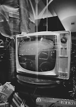 A vintage television set with dials sits on a table surrounded by patterned boxes. The reflection of nearby objects can be seen on the TV screen, and a jacket is visible next to it. The setting has a nostalgic, retro feel with a mix of geometric patterns.