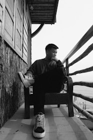 A person is sitting on a chair on a concrete patio beside a wooden structure with large windows. The person is dressed in casual clothing and sneakers, appearing to be in deep thought while looking away. The photo is taken in black and white, giving it a moody and contemplative vibe.