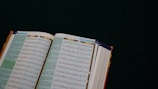 An open book with Arabic text on a dark green surface. The pages are filled with intricate script in black and colorful highlights, likely a religious or historical text.