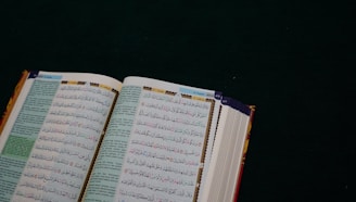 An open book with Arabic text on a dark green surface. The pages are filled with intricate script in black and colorful highlights, likely a religious or historical text.