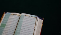 An open book with Arabic text on a dark green surface. The pages are filled with intricate script in black and colorful highlights, likely a religious or historical text.