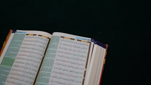 An open book with Arabic text on a dark green surface. The pages are filled with intricate script in black and colorful highlights, likely a religious or historical text.