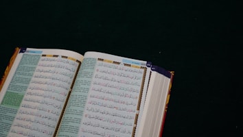 An open book with Arabic text on a dark green surface. The pages are filled with intricate script in black and colorful highlights, likely a religious or historical text.