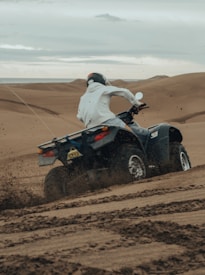 A person wearing a white hoodie and helmet is riding an ATV across a sandy desert landscape. The ATV kicks up dirt as it moves, capturing motion and the rugged terrain.