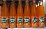 Several bottles of Green Spot California Orange soda are lined up on a shelf. The bottles are clear with an orange liquid inside, and each features a bright orange label with green and white text. The brand name 'Green Spot' is prominently displayed along with an illustration of an orange slice.