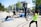 A group of people engaging in outdoor fitness activities in a park-like setting. They are using equipment such as a pull-up bar and battle ropes. The scene is sunny, with trees and modern buildings in the background.