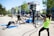 A group of people engaging in outdoor fitness activities in a park-like setting. They are using equipment such as a pull-up bar and battle ropes. The scene is sunny, with trees and modern buildings in the background.