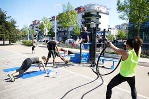 A group of people engaging in outdoor fitness activities in a park-like setting. They are using equipment such as a pull-up bar and battle ropes. The scene is sunny, with trees and modern buildings in the background.
