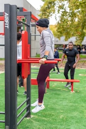 A person in athletic clothing is performing a dip exercise using outdoor gym equipment. The individual is dressed in a gray long-sleeve top, maroon leggings, and white sneakers. Several other people can be seen in the background, also engaged in fitness activities. The setting is an outdoor gym area with artificial grass and trees in the distance, creating a natural and active environment.