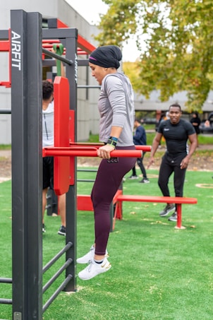 A person in athletic clothing is performing a dip exercise using outdoor gym equipment. The individual is dressed in a gray long-sleeve top, maroon leggings, and white sneakers. Several other people can be seen in the background, also engaged in fitness activities. The setting is an outdoor gym area with artificial grass and trees in the distance, creating a natural and active environment.