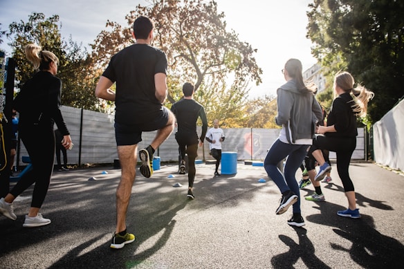 A group of people is engaging in an outdoor exercise session in a sunny environment. The participants appear to be jogging in place, with some wearing casual athletic clothing. The scene is set on a paved area surrounded by fencing and trees with autumn leaves.