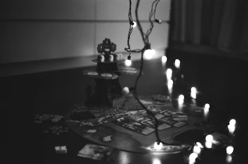 A black and white photograph of a board game setup with scattered pieces and cards. A set of string lights are draped above, casting small points of light across the scene. The game board features various details and sections, but the specific game is not clearly identifiable.