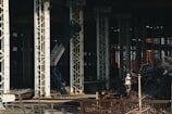 Large industrial warehouse or factory interior with tall metal pillars and overhead beams. A worker wearing a helmet is standing amidst a pile of scrap metal. The lighting is dim, creating a shadowy atmosphere.