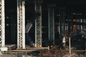 Large industrial warehouse or factory interior with tall metal pillars and overhead beams. A worker wearing a helmet is standing amidst a pile of scrap metal. The lighting is dim, creating a shadowy atmosphere.