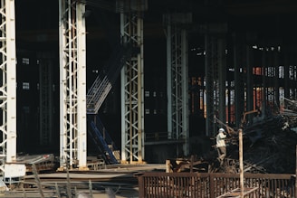 Large industrial warehouse or factory interior with tall metal pillars and overhead beams. A worker wearing a helmet is standing amidst a pile of scrap metal. The lighting is dim, creating a shadowy atmosphere.