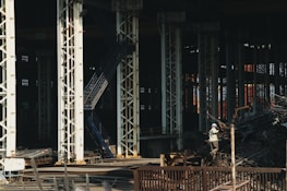 Large industrial warehouse or factory interior with tall metal pillars and overhead beams. A worker wearing a helmet is standing amidst a pile of scrap metal. The lighting is dim, creating a shadowy atmosphere.