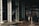 Large industrial warehouse or factory interior with tall metal pillars and overhead beams. A worker wearing a helmet is standing amidst a pile of scrap metal. The lighting is dim, creating a shadowy atmosphere.