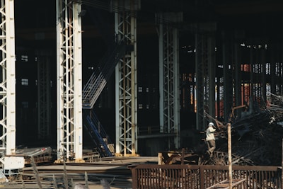 Large industrial warehouse or factory interior with tall metal pillars and overhead beams. A worker wearing a helmet is standing amidst a pile of scrap metal. The lighting is dim, creating a shadowy atmosphere.
