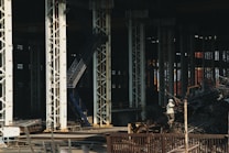 Large industrial warehouse or factory interior with tall metal pillars and overhead beams. A worker wearing a helmet is standing amidst a pile of scrap metal. The lighting is dim, creating a shadowy atmosphere.