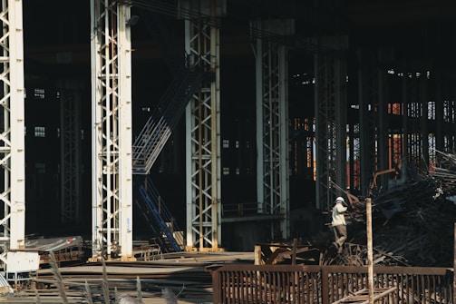 Large industrial warehouse or factory interior with tall metal pillars and overhead beams. A worker wearing a helmet is standing amidst a pile of scrap metal. The lighting is dim, creating a shadowy atmosphere.