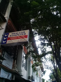 A sign for 'Shallom Komputer' is displayed prominently on the facade of a building, surrounded by lush green trees. The sign offers information about services related to hardware, software, networking, and accessories. The building features several rectangular windows and air conditioning units.