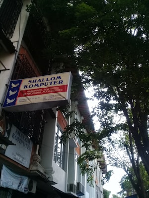 A sign for 'Shallom Komputer' is displayed prominently on the facade of a building, surrounded by lush green trees. The sign offers information about services related to hardware, software, networking, and accessories. The building features several rectangular windows and air conditioning units.