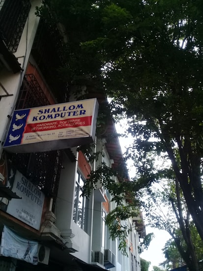 A sign for 'Shallom Komputer' is displayed prominently on the facade of a building, surrounded by lush green trees. The sign offers information about services related to hardware, software, networking, and accessories. The building features several rectangular windows and air conditioning units.