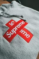 A gray hoodie is displayed with a prominent red and white logo. The logo consists of two intersecting rectangles, each with the word 'Supreme' in bold white letters.