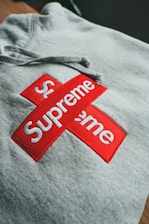 A gray hoodie is displayed with a prominent red and white logo. The logo consists of two intersecting rectangles, each with the word 'Supreme' in bold white letters.