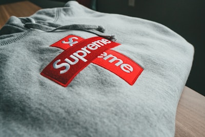 A folded gray hoodie features a prominent red and white cross logo, with the word 'Supreme' displayed prominently in bold white text.
