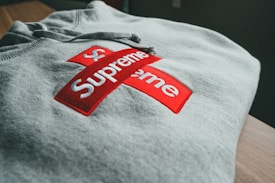 A folded gray hoodie features a prominent red and white cross logo, with the word 'Supreme' displayed prominently in bold white text.