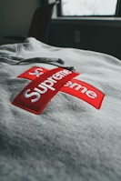 A close-up view of a gray sweatshirt featuring a red logo with white text in a cross shape, placed prominently on the fabric. The background shows a window with some light coming through, creating a soft, natural lighting effect on the garment.