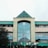 A large building with a pyramidal glass roof structure, displaying signs for Plaza Bogor. The facade appears weathered, with a blend of green and beige colors. A flagpole with a flag is positioned in front of the entrance. Trees partially obstruct the view on the left side.