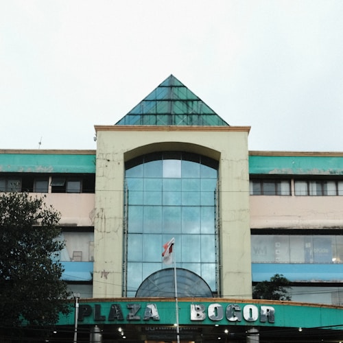 A large building with a pyramidal glass roof structure, displaying signs for Plaza Bogor. The facade appears weathered, with a blend of green and beige colors. A flagpole with a flag is positioned in front of the entrance. Trees partially obstruct the view on the left side.