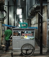 A small street food cart with a sign that reads 'PEMPEK' is situated in an alleyway with brick and concrete walls. The cart contains items for sale, and there is a chair on top. A blurred figure in a green shirt walks past, adding a sense of motion to the scene. Several illuminated signs hang above, providing light to the narrow passage.